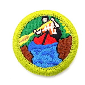 May include: Embroidered patch featuring a person in a boat, paddling on blue water. The patch has a green background with brown and red accents, and a yellow border. The person is wearing a white and orange shirt.