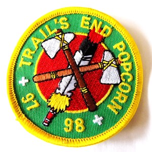 May include: A circular patch with a green background and yellow trim. The patch features the text "TRAIL'S END POPCORN" in yellow. The center has a red circle with crossed tomahawks and feathers. The numbers "97" and "98" are also present.