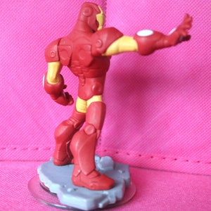 May include: A red and yellow Iron Man action figure, posed with one arm extended. The figure is on a grey base with a clear plastic disc. The figure's armour is red with yellow accents on the arms, legs, and head.