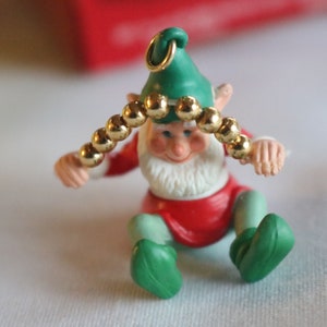 May include: A green and red Christmas ornament of a sitting elf wearing a green hat with a gold bead garland around his head.