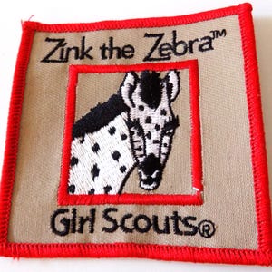 May include: A square patch with a red border and the words "Zink the Zebra" and "Girl Scouts" embroidered in black. The patch features a black and white zebra head inside a red square.