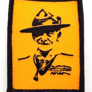 May include: A rectangular patch with a black border and a yellow background. The patch features a black silhouette of a man wearing a hat and a neckerchief. The man's signature is at the bottom.