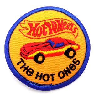 May include: Embroidered patch with a yellow background and a blue border. The patch features a red car graphic and the words "Hot Wheels" in red script above the car. Below the car, the text reads "The Hot Ones" in black.