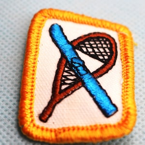 May include: A yellow embroidered patch with a blue and brown lacrosse stick and net.