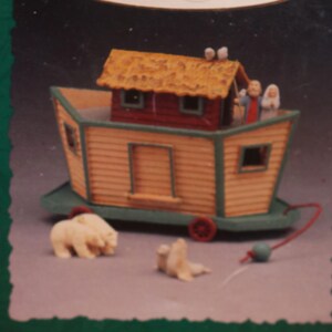 May include: Keepsake ornament featuring a miniature ark with a tan exterior, red roof, and green trim. The ark has small figures on top and includes miniature polar bears and a seal. The words "Keepsake Ornament" are at the top.