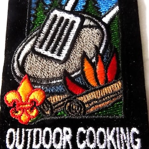 May include: A rectangular black embroidered patch featuring a scout fleur-de-lis emblem in yellow and red, a gray spatula over a frying pan, and a campfire with orange and red flames. The text OUTDOOR COOKING is stitched in white at the bottom.