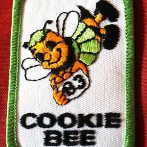 May include: Green and white embroidered patch with a cartoon bee holding a box with the number 83 on it. The text "COOKIE BEE" is below the bee.