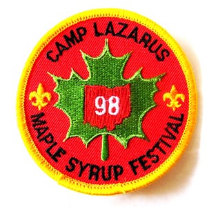 May include: A round, embroidered patch with a red background and yellow border. The patch features the text "CAMP LAZARUS MAPLE SYRUP FESTIVAL" in black. A green maple leaf design with a red square containing the number "98" is in the center.