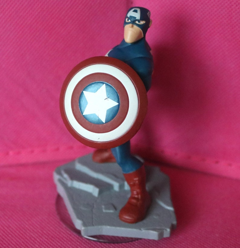 Puede incluir: Una figura de pl&aacute;stico de Capit&aacute;n Am&eacute;rica sosteniendo su escudo. El escudo es rojo, blanco y azul con una estrella blanca en el centro. La figura est&aacute; de pie sobre una base gris.