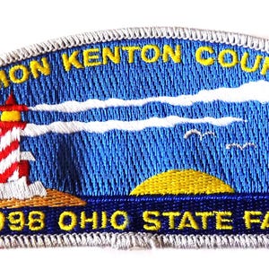 May include: Embroidered patch featuring a lighthouse with red and white stripes, a yellow sun, and the text "SIMON KENTON COUNCIL" and "1998 OHIO STATE FAIR". The patch has a blue background with white clouds and a fleur-de-lis.
