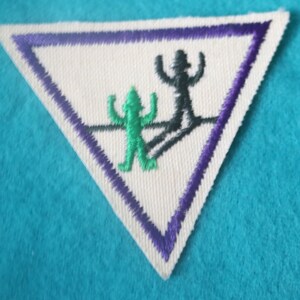 May include: A triangular shaped patch with a purple border. The patch features a black silhouette of a person with arms raised, and a green silhouette of a person with arms raised. The green silhouette is standing on a line, and the black silhouette is standing on a line above the green silhouette.