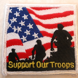May include: An embroidered patch with a red, white, and blue American flag and three silhouetted figures standing in front of it. The text "Support Our Troops" is written in yellow below the figures.