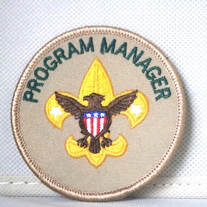 May include: A tan round embroidered patch with a green border. The patch features the Boy Scouts of America logo with the words "PROGRAM MANAGER" around the outside.