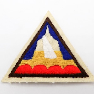 May include: Embroidered patch in a triangular shape. The patch features a design with a white central element, blue and yellow sections, and a red base. The border is brown, and the background is beige.