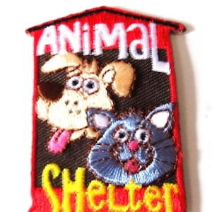 May include: Embroidered patch featuring a dog and cat with the words "ANIMAL Shelter". The patch has a red border and a black background. The dog is tan with brown spots and a pink tongue. The cat is blue with pink accents.