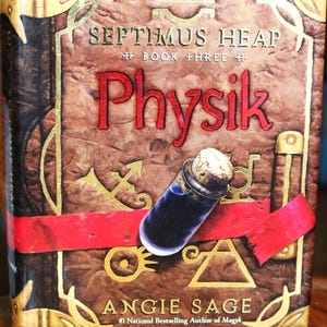 May include: Book cover for "Septimus Heap: Physik", book three in the series. The cover features a red ribbon tied around a brown leather-bound book with gold accents. The title "Physik" is in red lettering and the author's name, "Angie Sage", is below the title.