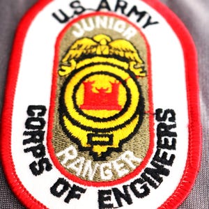 May include: A red, white, and black oval patch with the text "US Army Junior Corps of Engineers Ranger" embroidered on it.