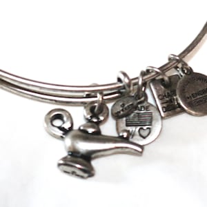 May include: Silver-toned bangle bracelet with multiple charms. Charms include a genie lamp, a small tag with a heart and American flag, and two round charms with text. The bracelet is on a white background.