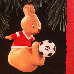 May include: A brown and tan kangaroo wearing a red and white jersey kicks a black and white soccer ball with the number "94" on it. The ornament is labeled "Kickin' Roo".