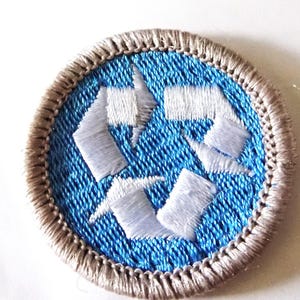 May include: Circular embroidered patch featuring the recycling symbol. The symbol is white, set against a textured blue background, and framed by a tan border. The patch is designed for environmental awareness and promoting recycling.