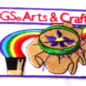 May include: Embroidered patch with the text "GS Arts & Crafts" above a design featuring a rainbow, a hand holding a green and purple emblem, and a black pot. The patch has a white background and a purple border.