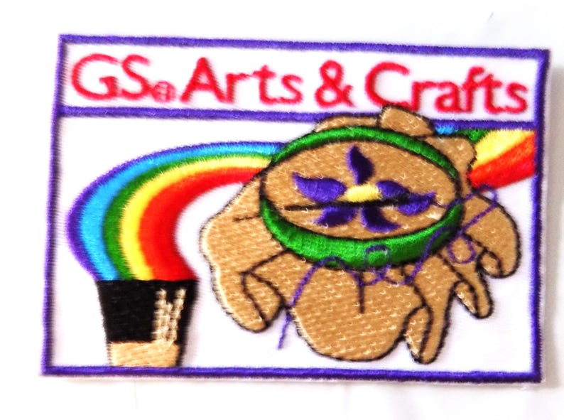 May include: Embroidered patch with the text "GS Arts & Crafts" above a design featuring a rainbow, a hand holding a green and purple emblem, and a black pot. The patch has a white background and a purple border.