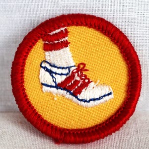 May include: A red, white, and blue embroidered patch with a yellow background. The patch features a white shoe with red and blue accents.