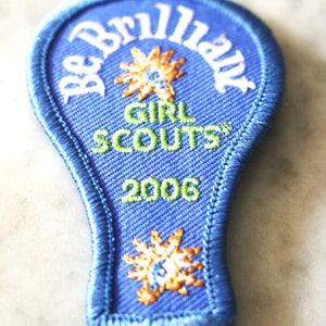 May include: A blue lightbulb-shaped Girl Scout patch with the text "Be Brilliant" in white, "Girl Scouts" in green and white, and "2006" in green. The patch features two yellow and orange embroidered stars.