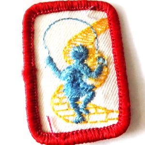 May include: A rectangular embroidered patch with a red stitched border on a white background. The design features a blue figure jumping rope, positioned over a yellow brick path pattern. The embroidery uses blue, yellow, and red thread.
