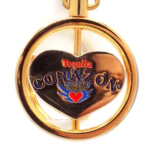 May include: A gold-colored metal pendant with a heart-shaped center. The heart features the words "Tequila Corazon de Agave" in red and blue, with a red heart and blue wings design. The pendant is circular.