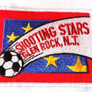May include: Embroidered patch with a soccer ball and the words "SHOOTING STARS GLEN ROCK, N.J." on a white banner. The patch has a red border, a blue background with yellow stars, and a white stitched edge.