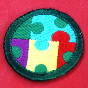 Pode incluir: Um remendo redondo de tecido verde com um design de pe&ccedil;as de quebra-cabe&ccedil;a coloridas. As pe&ccedil;as do quebra-cabe&ccedil;a s&atilde;o em tons de azul, vermelho, amarelo, roxo e verde. O remendo tem uma borda verde.