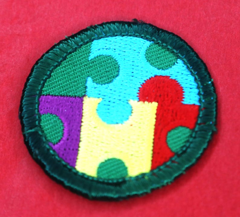 Pode incluir: Um remendo redondo de tecido verde com um design de pe&ccedil;as de quebra-cabe&ccedil;a coloridas. As pe&ccedil;as do quebra-cabe&ccedil;a s&atilde;o em tons de azul, vermelho, amarelo, roxo e verde. O remendo tem uma borda verde.