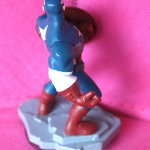 Puede incluir: Una figura de acci&oacute;n de pl&aacute;stico azul y rojo de Capit&aacute;n Am&eacute;rica de pie sobre una base gris. Sostiene un escudo rojo, blanco y azul en su mano derecha.