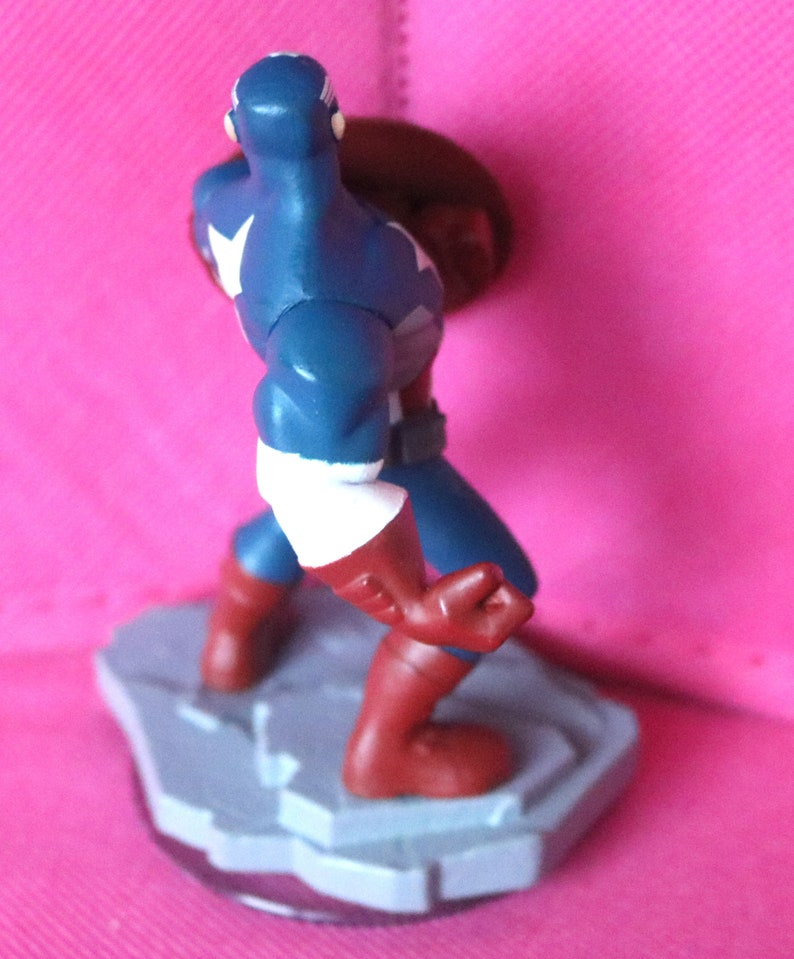 Puede incluir: Una figura de acci&oacute;n de pl&aacute;stico azul y rojo de Capit&aacute;n Am&eacute;rica de pie sobre una base gris. Sostiene un escudo rojo, blanco y azul en su mano derecha.