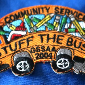 May include: Orange school bus patch with the words "Community Service Stuff the Bus" and "GSSAA 2004". The bus is filled with colorful pencils and other school supplies.