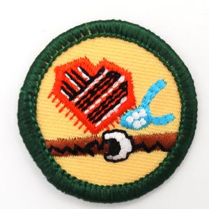 May include: A round embroidered patch with a yellow background, featuring a red and black heart-shaped design, a blue object, and a brown belt with a white buckle. The patch has a green border.