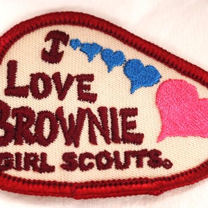 May include: Embroidered patch with a cream-colored background and a red border. The patch features the words "I LOVE BROWNIE GIRL SCOUTS" in maroon thread, along with blue and pink heart shapes. A vintage item.