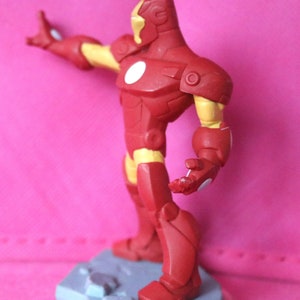 May include: A red and yellow Iron Man action figure, posed with one arm extended. The figure has a white circle on its chest and stands on a grey base. The figure's design is based on the Marvel Comics character.
