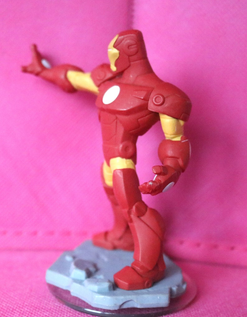 May include: A red and yellow Iron Man action figure, posed with one arm extended. The figure has a white circle on its chest and stands on a grey base. The figure's design is based on the Marvel Comics character.
