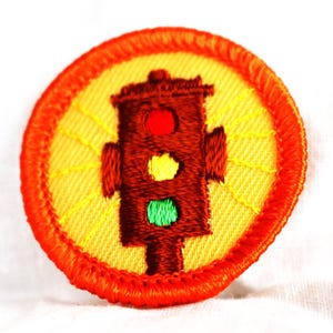 May include: A round, orange-bordered patch with a yellow background. A brown traffic light with red, yellow, and green lights is in the center of the patch. The traffic light is surrounded by yellow rays of light.
