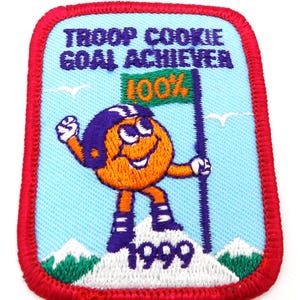 May include: Embroidered patch with a red border, featuring a cartoon character holding a flag that reads "100%". The patch says "TROOP COOKIE GOAL ACHIEVER" and "1999". The character is orange with a purple and white helmet, standing on a snowy mountain.