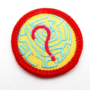 May include: A round embroidered patch with a yellow center featuring a blue maze design and a red question mark. The patch has a red border. This patch could be used on clothing or accessories.
