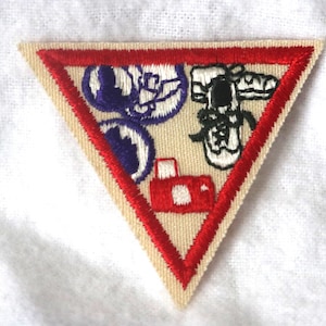 May include: A triangular scout patch with a red border. The patch features a camera, a pair of shoes, and a purple and white design.