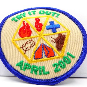 May include: Embroidered patch with a beige background and a blue border. The patch features a yellow geometric design with various symbols, including a flame, musical note, cross, bear, rope, and tent. The text "TRY IT OUT!" and "APRIL 2001" are embroidered in green.