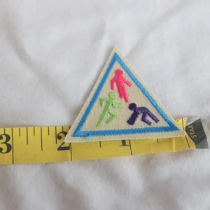 May include: A triangular Girl Scout patch with a blue border. The patch features three figures in a triangle, one pink, one green, and one purple. The figures are running and walking.