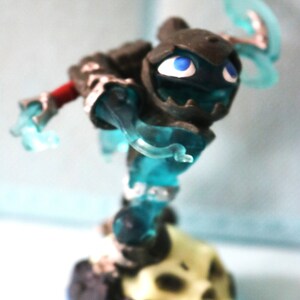 May include: A blue and gray action figure of a character with a skull base. The character has a blue translucent body and a gray head with blue eyes.