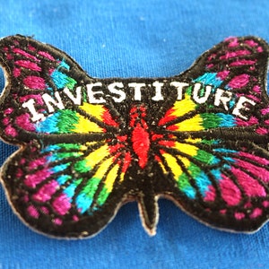 May include: A colorful butterfly-shaped patch with the word "INVESTITURE" embroidered in white on a black background. The butterfly has rainbow wings with shades of red, orange, yellow, green, blue, and purple.