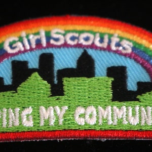 May include: A rainbow-colored embroidered patch with the text "Girl Scouts" and "Helping My Community". The patch features a cityscape with a green hill in the foreground.