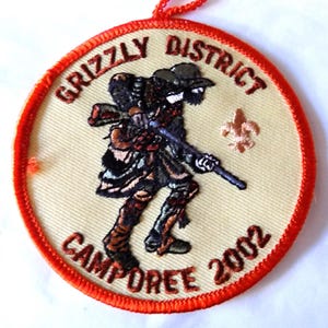 May include: Embroidered patch with a red border and the words "GRIZZLY DISTRICT CAMPOREE 2002." The patch features a detailed illustration of a person in a hat and carrying a rifle. A fleur-de-lis is also present.